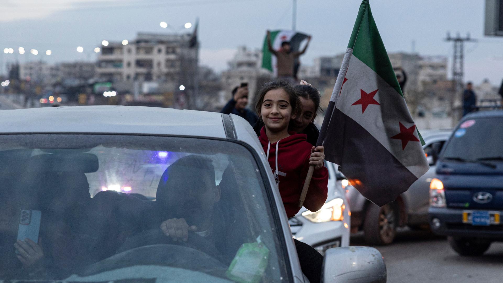 Autos stehen im Stau auf einer Straße. Aus einem Autofenster reckt sich ein Mädchen hervor und hält die Flagge der syrischen Opposition.