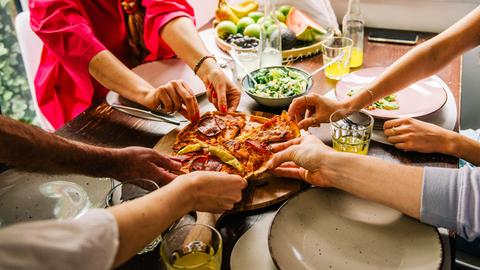 An einem Tisch sitzende Personen greifen nach den Stücken einer Pizza.