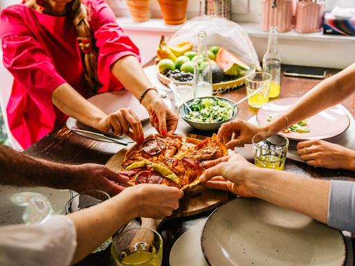 An einem Tisch sitzende Personen greifen nach den Stücken einer Pizza.