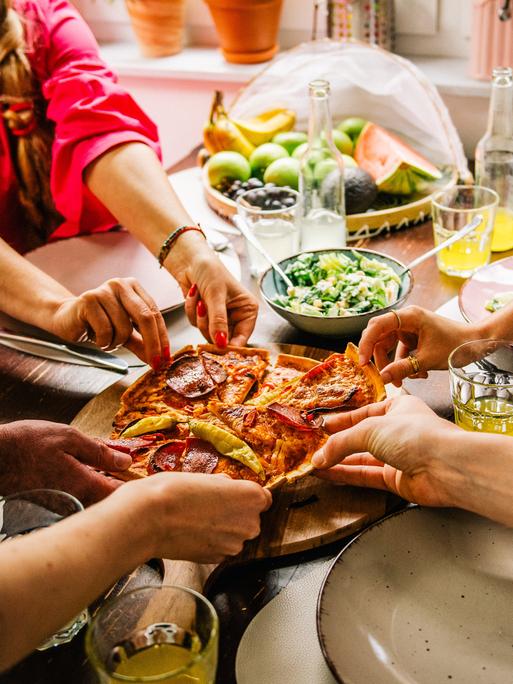An einem Tisch sitzende Personen greifen nach den Stücken einer Pizza.