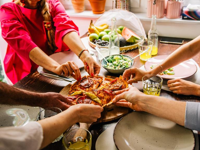 An einem Tisch sitzende Personen greifen nach den Stücken einer Pizza.