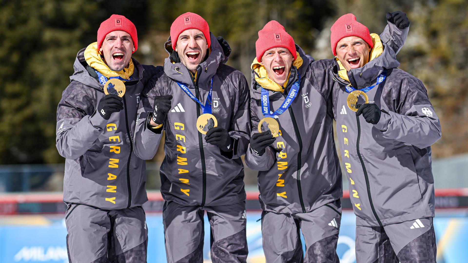 Die vier Biathleten stehen nebeneinander, halten ihre Goldmedaillen hoch und jubeln. Sie tragen rote Mützen und dunkelgraue Skianzüge. Die vier Biathleten stehen nebeneinander, halten ihre Goldmedaillen hoch und jubeln. Sie tragen rote Mützen und dunkelgraue Skianzüge.