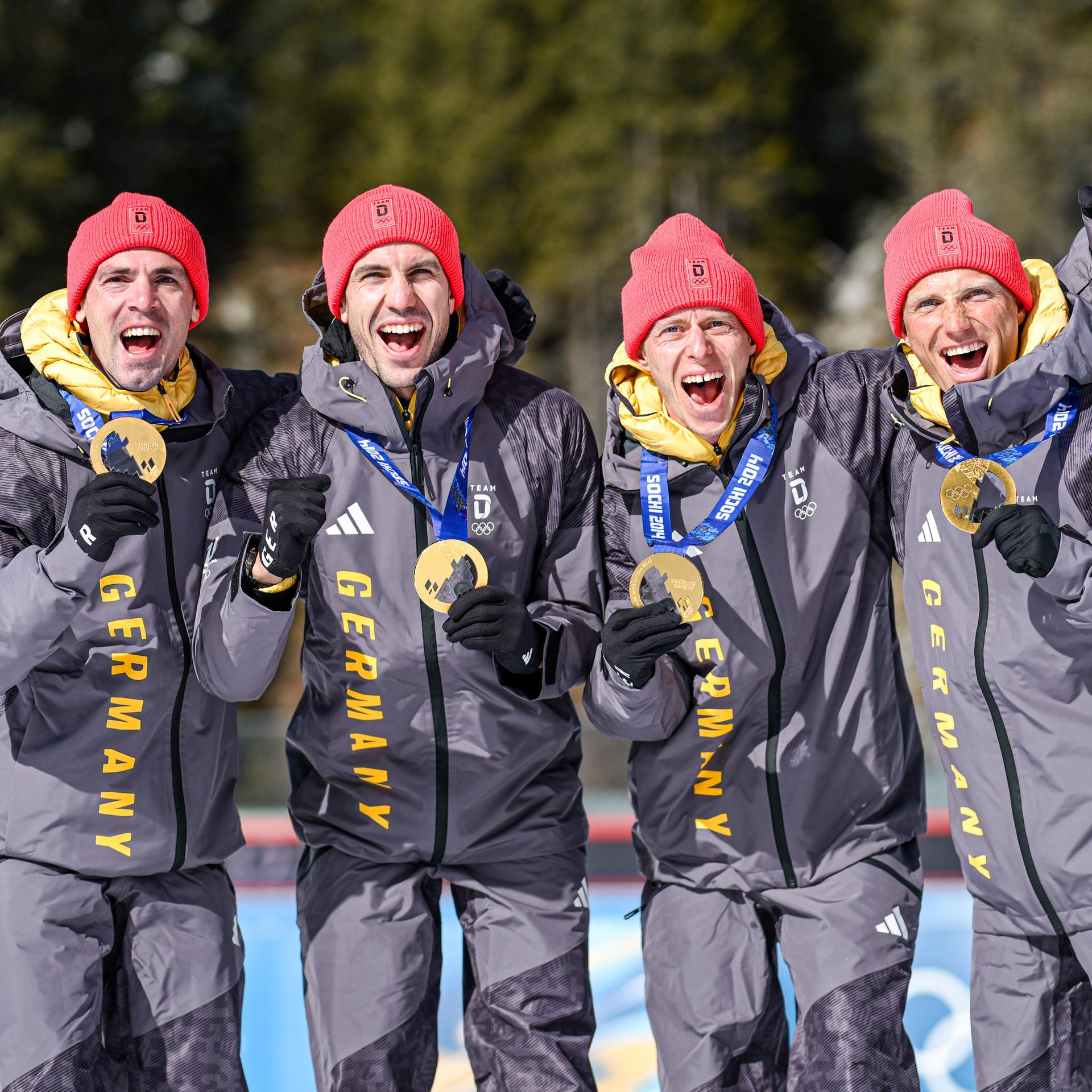 Die vier stehen nebeneinander, halten ihre Goldmedaillen hoch und jubeln. Sie tragen rote Mützen und dunkelgraue Skianzüge. 