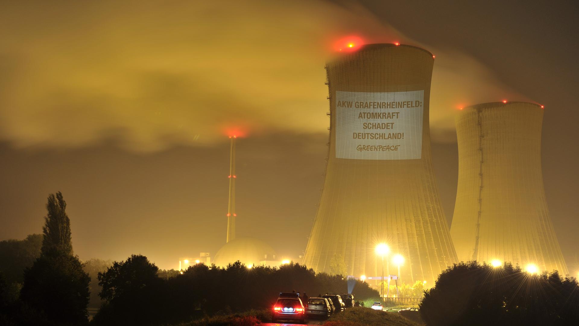 Ein Kühlturm eines Atomkraftwerks ist bei Dunkelheit beleuchtet. An dem Turm hängt ein Banner einer Greenpeace-Aktion mit der Aufschrift "AKW Grafenrheinfeld - Atomkraft schadet Deutschland".