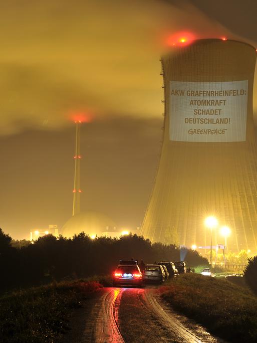 Ein Kühlturm eines Atomkraftwerks ist bei Dunkelheit beleuchtet. An dem Turm hängt ein Banner einer Greenpeace-Aktion mit der Aufschrift "AKW Grafenrheinfeld - Atomkraft schadet Deutschland".