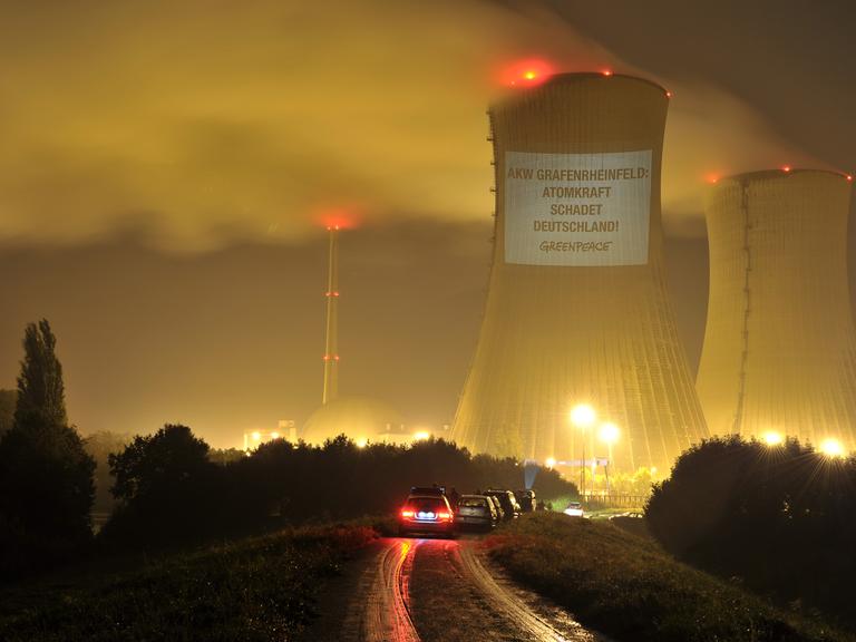 Ein Kühlturm eines Atomkraftwerks ist bei Dunkelheit beleuchtet. An dem Turm hängt ein Banner einer Greenpeace-Aktion mit der Aufschrift "AKW Grafenrheinfeld - Atomkraft schadet Deutschland".