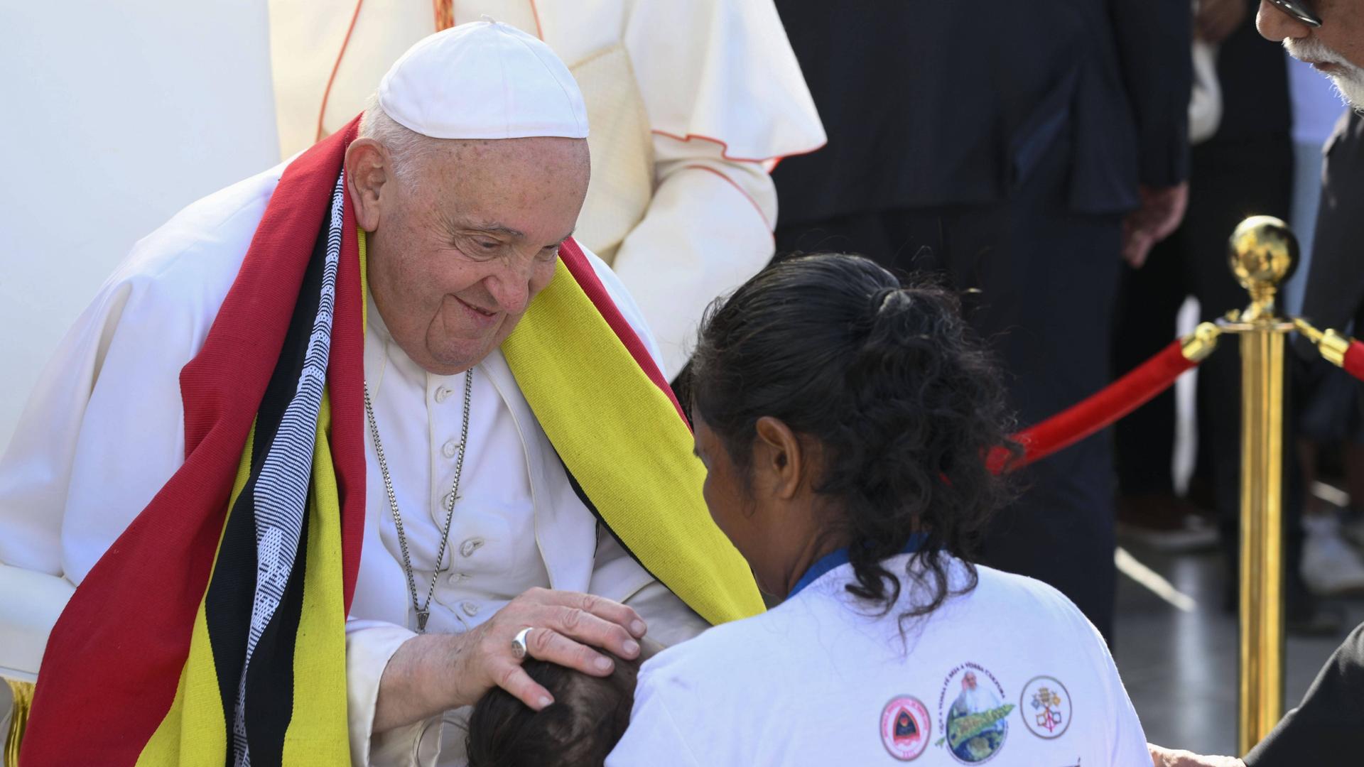 Papst Franziskus legt einem Kind die Hand auf den Kopf, das von einer Frau gehalten wird.