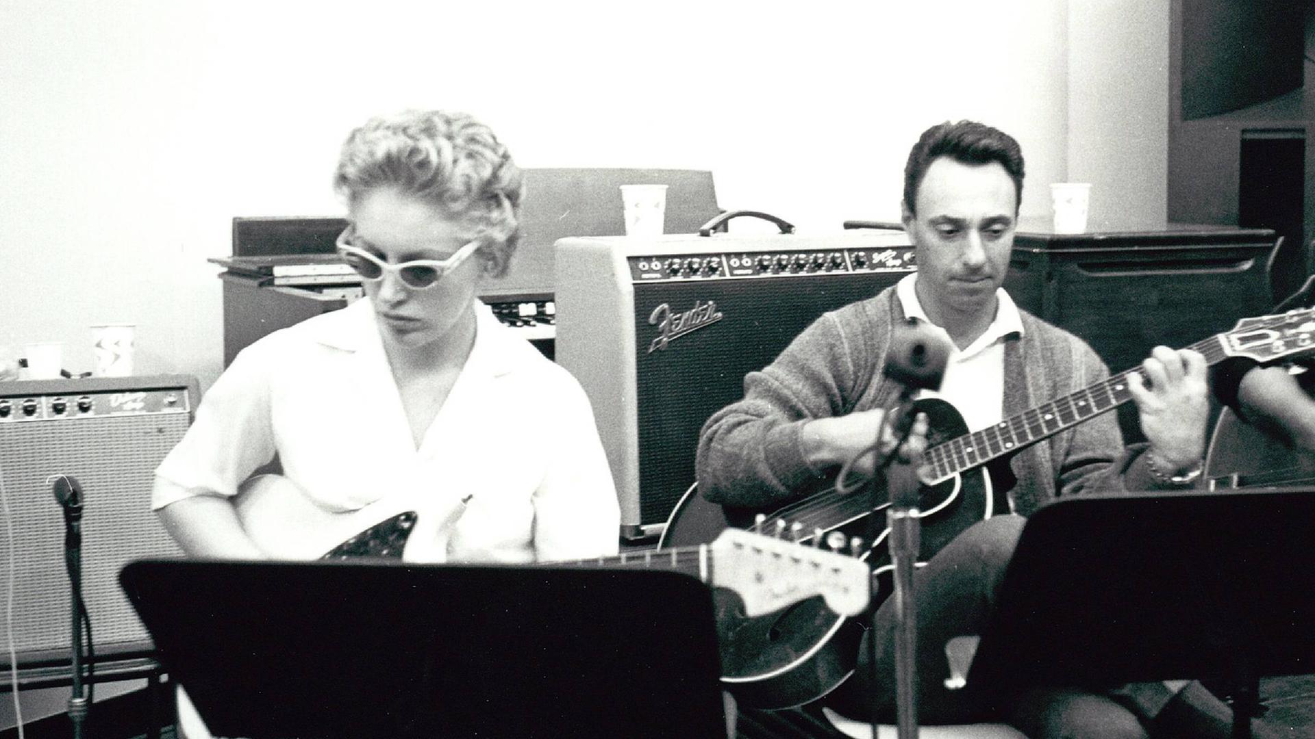 Auf dem schwarz-weiß Bild steht Carol Kaye hinter einem Notenständer mit ihrem Bass im Studio, daneben ein weiterer Musiker Auf dem schwarz-weiß Bild steht Carol Kaye hinter einem Notenständer mit ihrem Bass im Studio, daneben ein weiterer Musiker