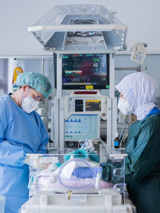 Ein Arzt und eine Krankenschwester stehen in Schutzanzug mit Mundschutz an einem Intensivbett auf der Neugeborenenstation.
