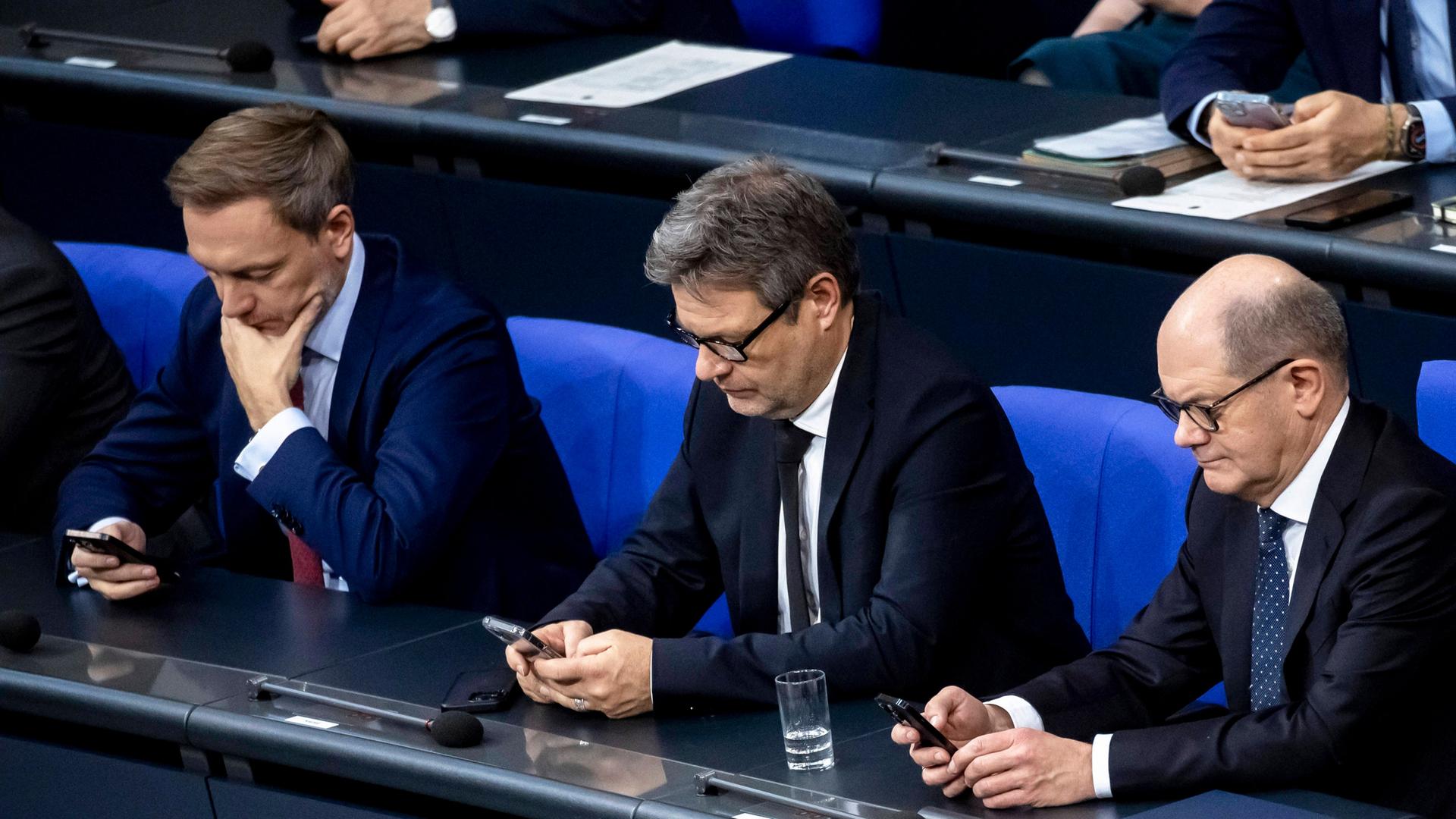 Bundeskanzler Olaf Scholz, Bundeswirtschaftsminister Robert Habeck und Bundesfinanzminister Christian Lindner sitzen in der Regierungsbank im Bundestag und blicken auf ihre Handys.