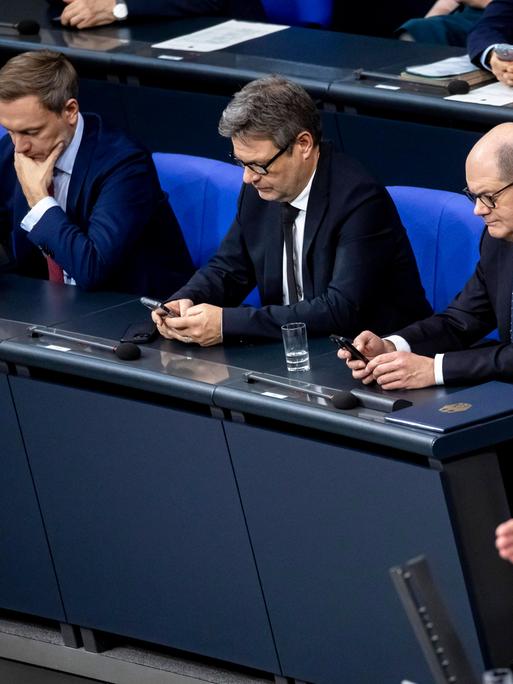 Bundeskanzler Olaf Scholz, Bundeswirtschaftsminister Robert Habeck und Bundesfinanzminister Christian Lindner sitzen in der Regierungsbank im Bundestag und blicken auf ihre Handys.