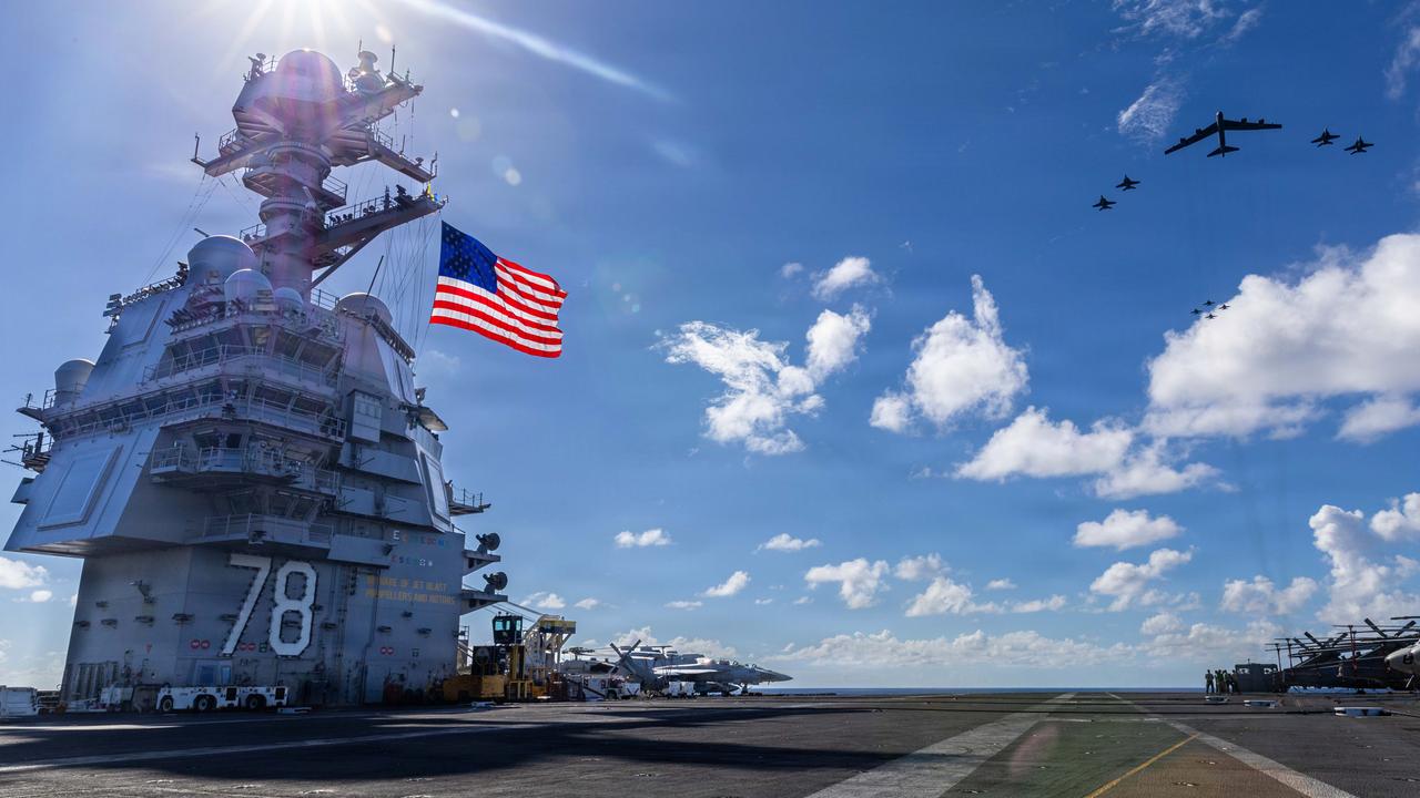 Blick über den Flugzeugträger, links im Bild die US-Flagge an einem turmartigen Aufbau, rechts vor blauem Himmel Kampfjets.