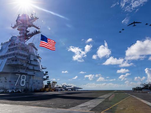 Blick über den Flugzeugträger, links im Bild die US-Flagge an einem turmartigen Aufbau, rechts vor blauem Himmel Kampfjets.