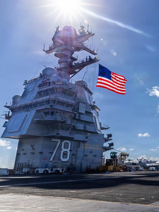 Blick über den Flugzeugträger, links im Bild die US-Flagge an einem turmartigen Aufbau, rechts vor blauem Himmel Kampfjets.
