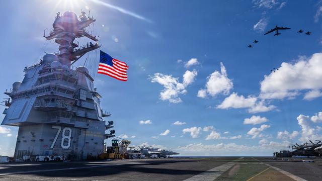Blick über den Flugzeugträger, links im Bild die US-Flagge an einem turmartigen Aufbau, rechts vor blauem Himmel Kampfjets.