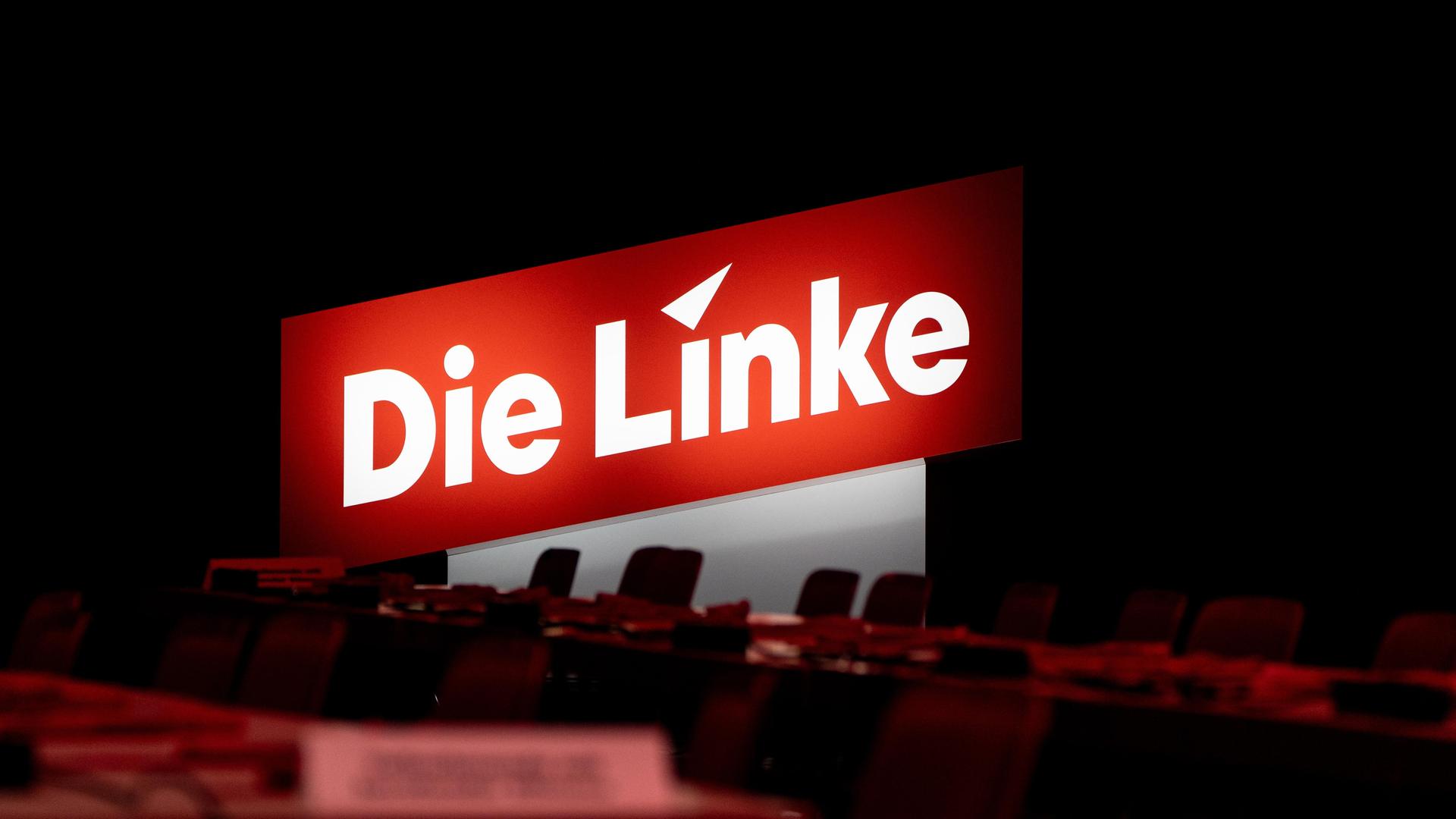 Parteilogo von  Die Linke  hinter leeren Stühlen in einem Saal.