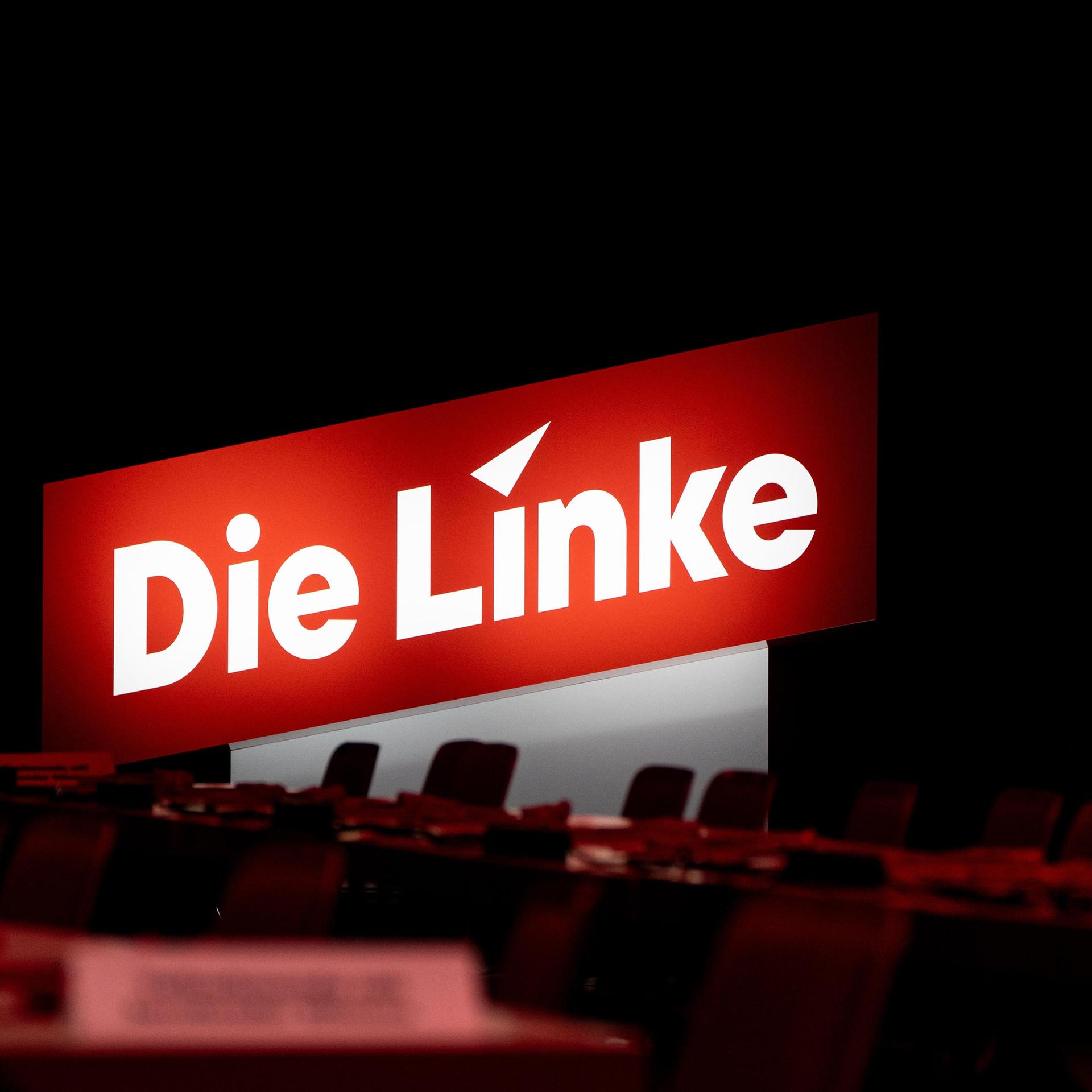 Parteilogo von  Die Linke  hinter leeren Stühlen in einem Saal.