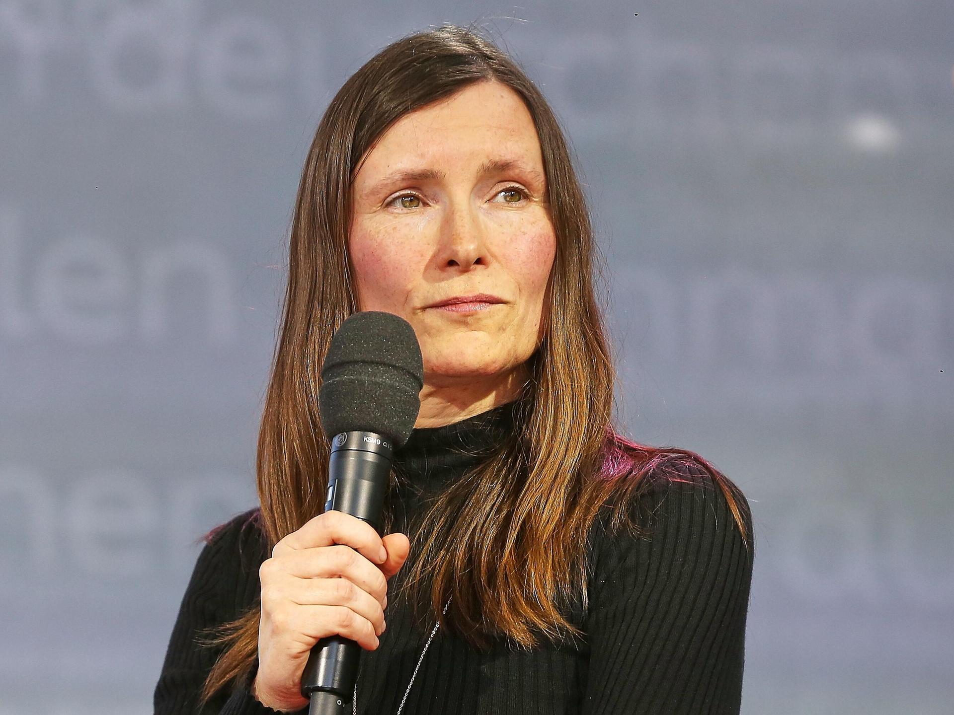 Heike Geißler auf der Leipziger Buchmesse. Sie trägt ein schwarzes Oberteil und hällt ein Mikrofon in der Hand. Der Hintergrund ist grau.