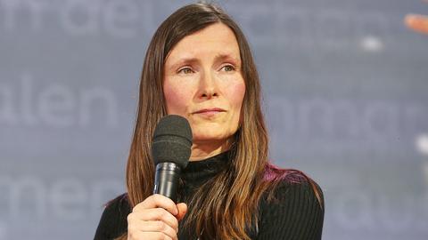 Heike Geißler auf der Leipziger Buchmesse. Sie trägt ein schwarzes Oberteil und hällt ein Mikrofon in der Hand. Der Hintergrund ist grau.