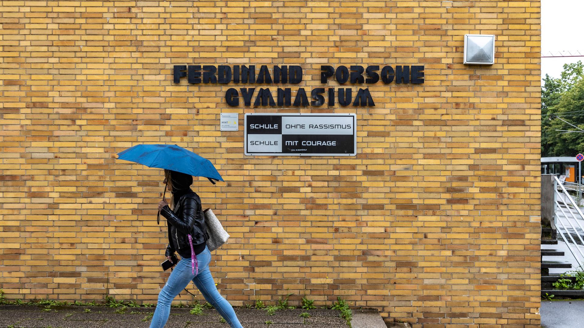 Eine Schülerin läuft mit gespanntem Schirm unter dem Namensschild des "Ferdinand Porsche Gymnasiums". Unter dem Namen steht "Schule ohne Rassismus".