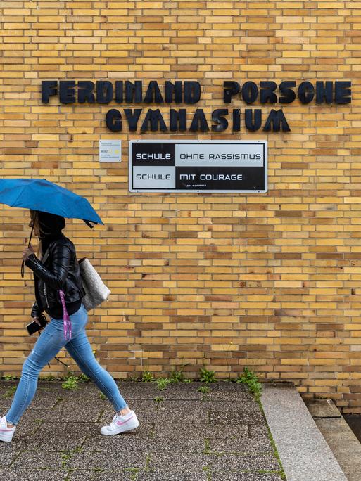 Eine Schülerin läuft mit gespanntem Schirm unter dem Namensschild des "Ferdinand Porsche Gymnasiums". Unter dem Namen steht "Schule ohne Rassismus".