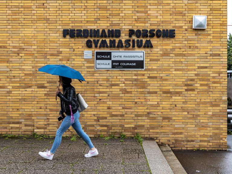 Eine Schülerin läuft mit gespanntem Schirm unter dem Namensschild des "Ferdinand Porsche Gymnasiums". Unter dem Namen steht "Schule ohne Rassismus".