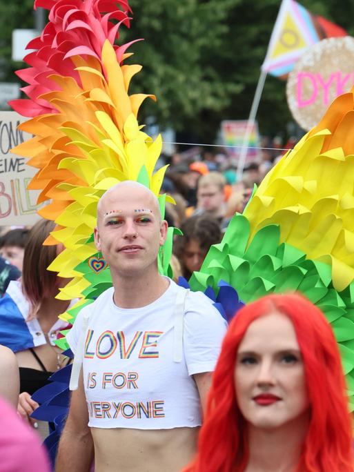 Menschen bei der CSD Demonstration in Berlin. Im Vordergrund eine Frau mit roten Haaren und ein Mann mit Flügeln in Regenbogenfarben. 