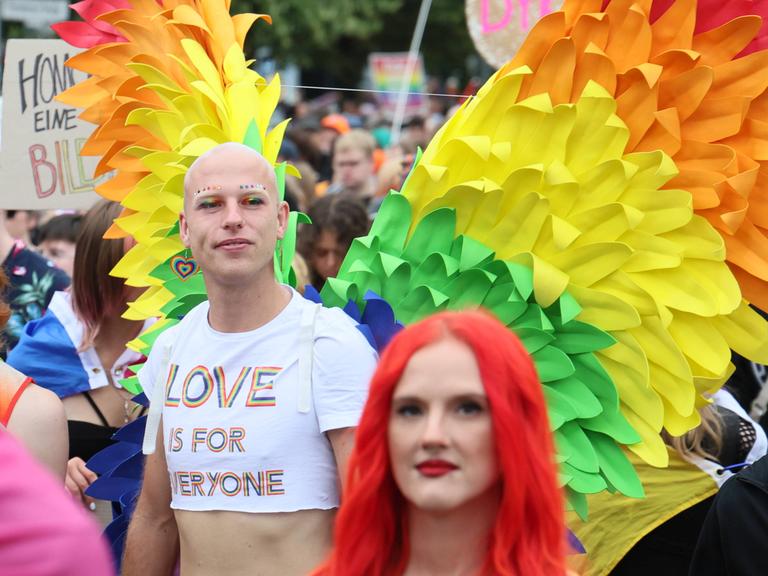 Menschen bei der CSD Demonstration in Berlin. Im Vordergrund eine Frau mit roten Haaren und ein Mann mit Flügeln in Regenbogenfarben. 
