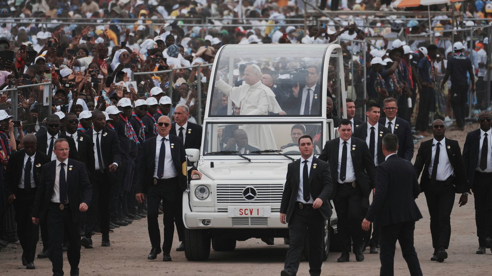Papst Leo XIV. kommt in Kilamba an, einem Vorort der Hauptstadt Luanda. Er steht in einem Fahrzeug und begrüßt die Gläubigen. Leibwächter sichern das Auto.