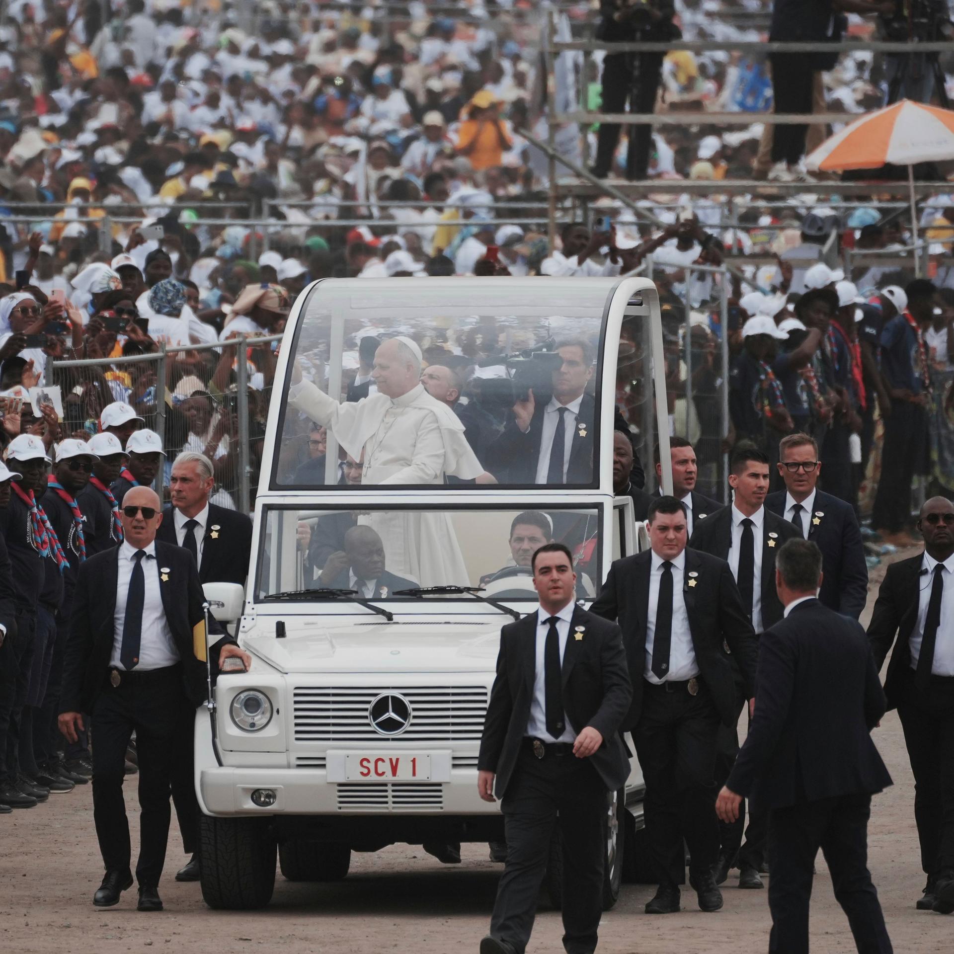 Papst Leo XIV. kommt in Kilamba, einem Vorort der Hauptstadt Luanda. Er steht in einem Fahrzeug und begrüßt die Gläubigen. Leibwächter sichern das Auto.