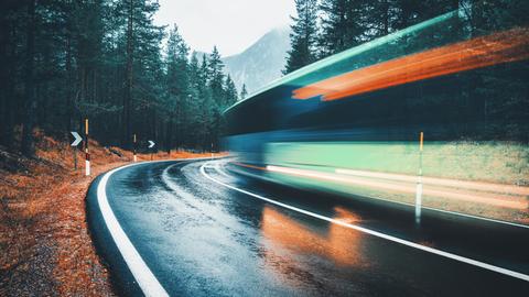 Blick auf eine Straße im Wald in dynamischer Perspektive: Ein Bus fährt die Straße entlang, man erkennt von ihm allerdings nur Schlieren.