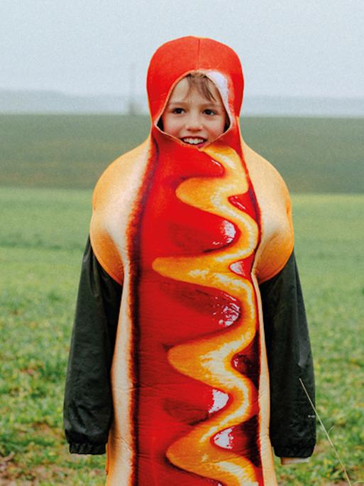 Ein kleiner Junge steht in einem Hotdog-Kostüm auf einem grünen Feld.