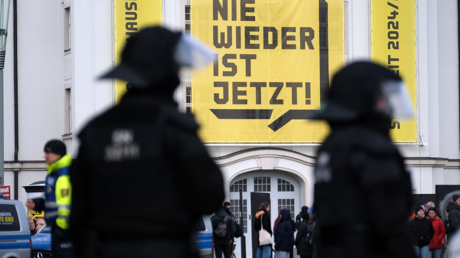 Polizisten am Rande einer Kundgebung in Dresden. Im Hintergrund ist ein Plakat mit der Aufschrift "Nie wieder ist jetzt!" zu sehen. Polizisten am Rande einer Kundgebung in Dresden. Im Hintergrund ist ein Plakat mit der Aufschrift "Nie wieder ist jetzt!" zu sehen.