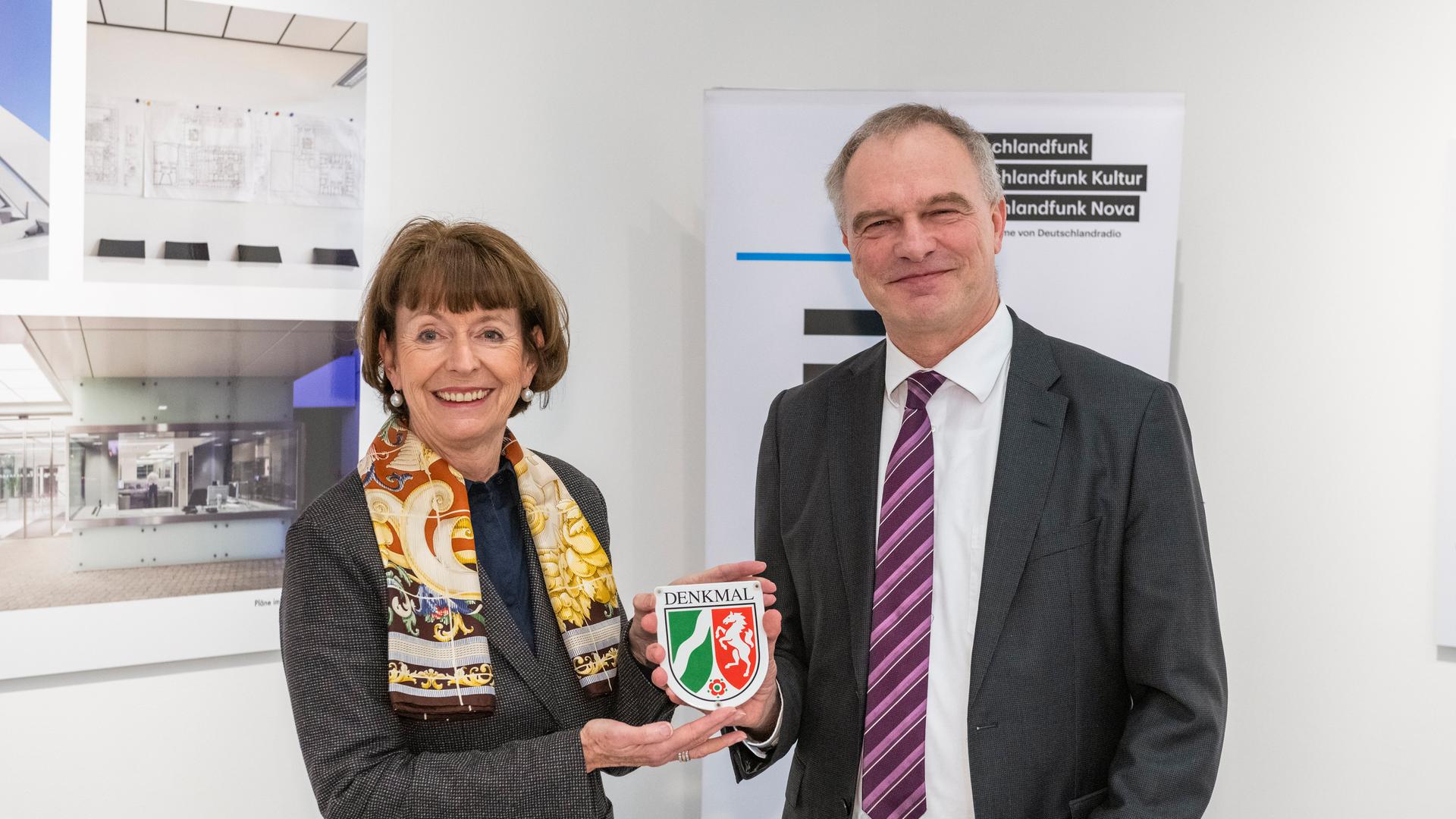 Die Oberbürgermeisterin Henriette Reker und Deutschlandradio-Intendant Stefan Raue halten gemeinsam die NRW-Plakette für den Denkmalschutz des Deutschlandfunk-Gebäudes. 