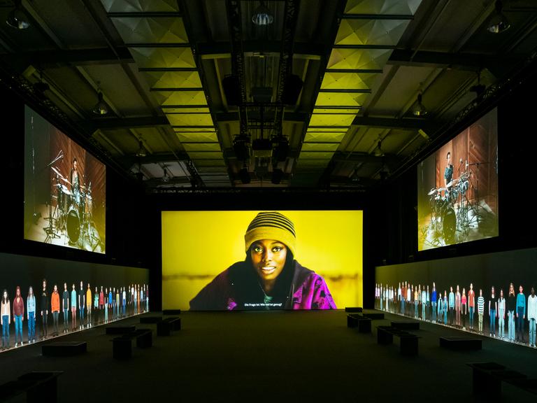 In einer großen Halle werden Bilder auf fünf Bildschirme projiziert. Das zentralste Bild zeigt die Nahaufnahme einer Schwarzen Frau. Auf den seitlichen Monitoren sind Schlagzeugspieler und nebeneinander stehende Menschen in Lebensgröße zu sehen.