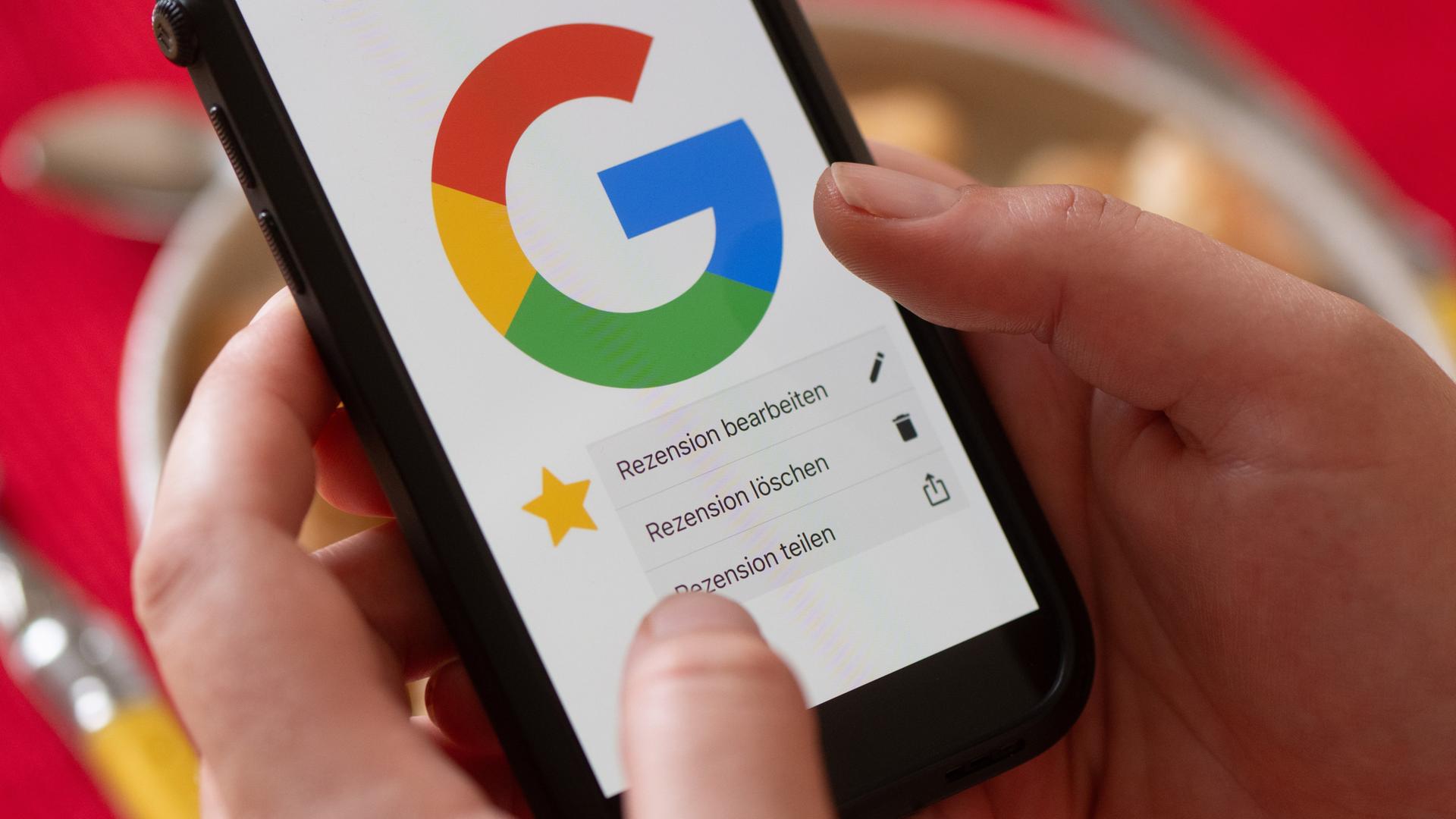 Eine Frau hält vor einem gedeckten Esstisch ein Smartphone mit dem Google-Logo und dem Menüpunkt eine Rezension zu bearbeiten, zu löschen oder zu teilen. Eine Frau hält vor einem gedeckten Esstisch ein Smartphone mit dem Google-Logo und dem Menüpunkt eine Rezension zu bearbeiten, zu löschen oder zu teilen.