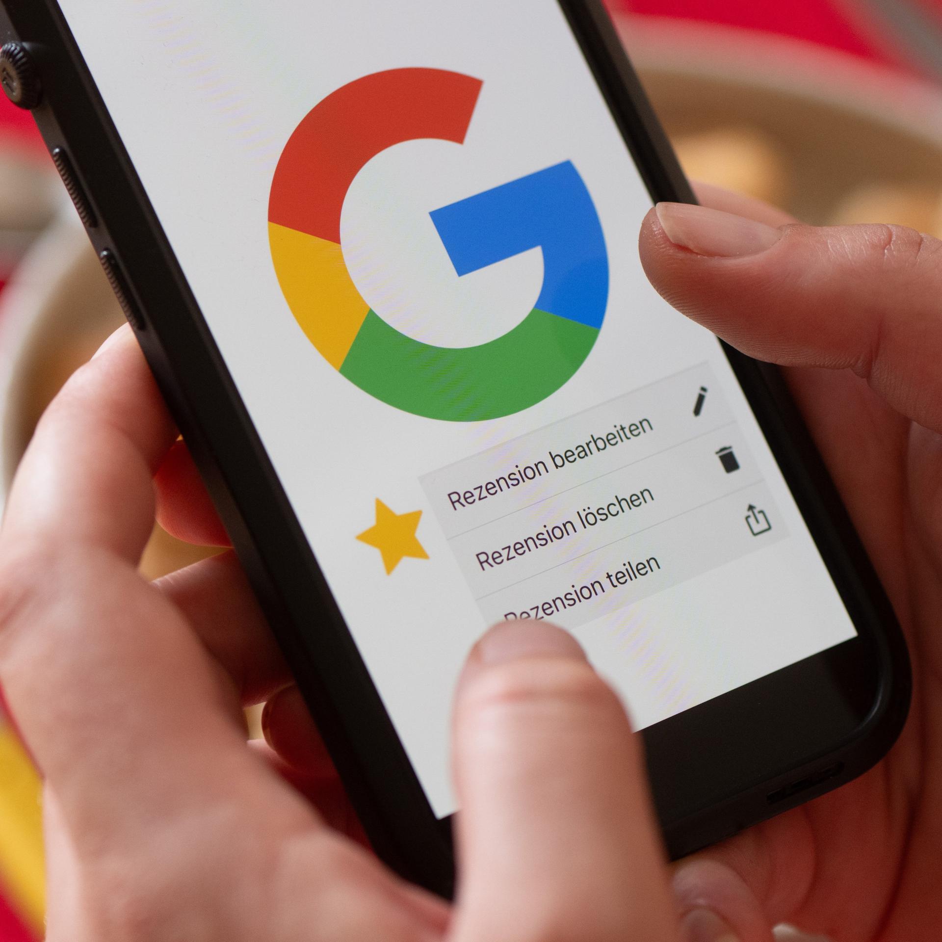 Eine Frau hält vor einem gedeckten Esstisch ein Smartphone mit dem Google-Logo und dem Menüpunkt eine Rezension zu bearbeiten, zu löschen oder zu teilen. 