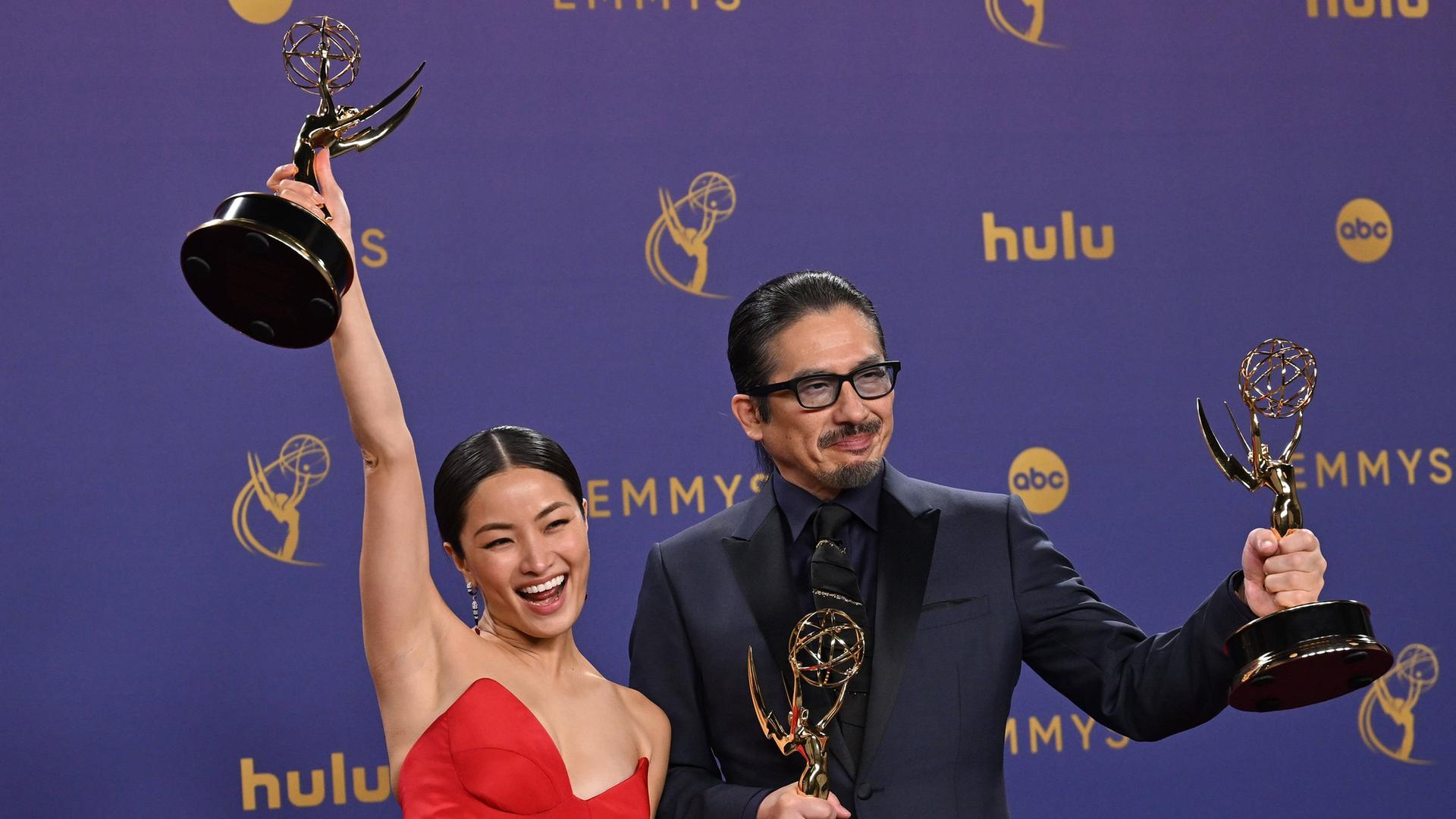 Die Schauspieler Anna Sawai und Hiroyuki Sanada halten Emmy-Preise hoch und lachen. Sie stehen nebeneinander. Sie trägt ein rotes Kleid. Er trägt einen Anzug.