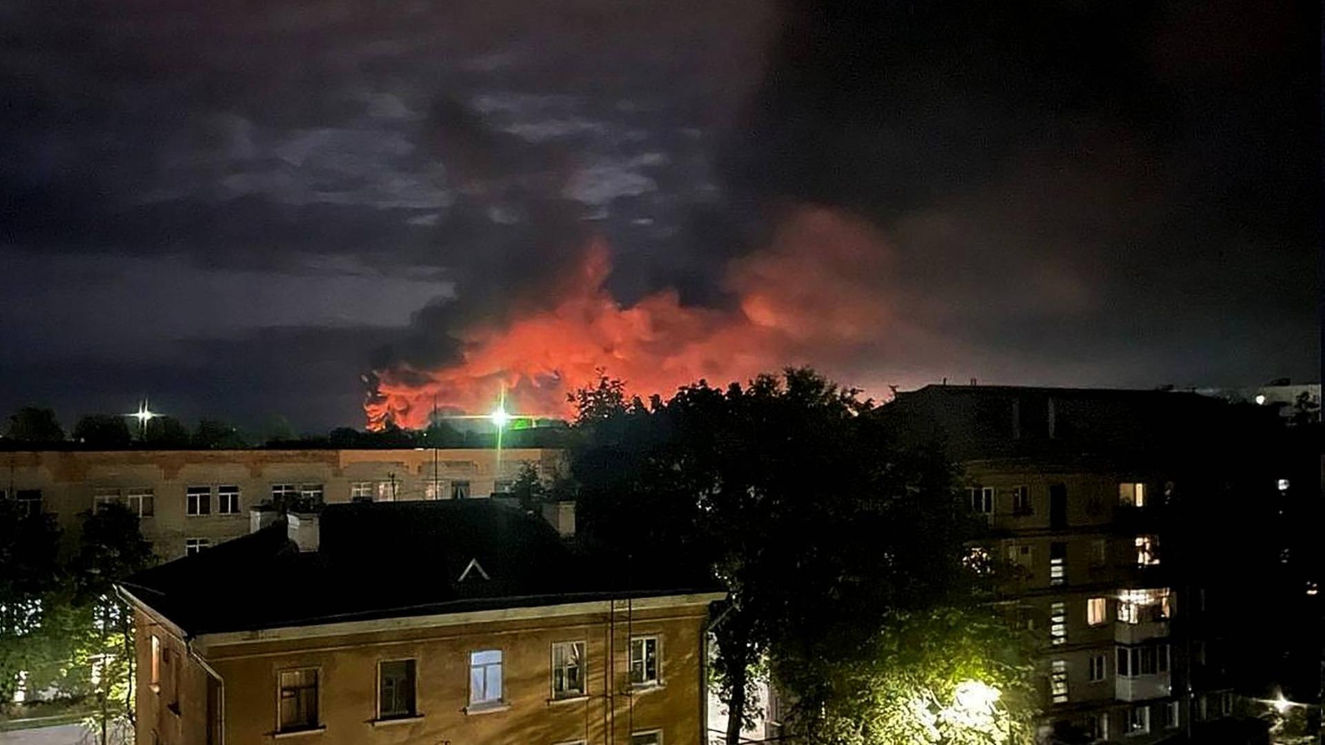 Nachtaufnahme einer Stadt. Im Hintergrund steigen rote Rauchwolken auf.