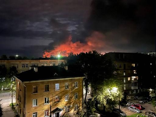 Nachtaufnahme einer Stadt. Im Hintergrund steigen rote Rauchwolken auf.