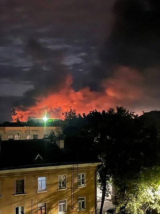 Nachtaufnahme einer Stadt. Im Hintergrund steigen rote Rauchwolken auf.