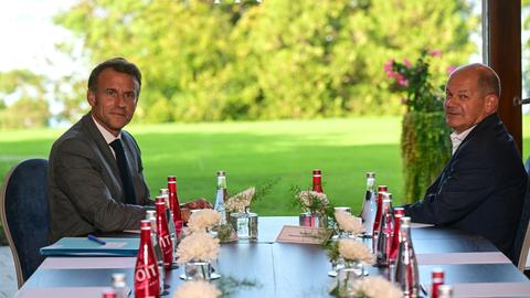 Macron und Scholz sitzen sich an einem Tisch gegenüber und schauen seitlich in Richtung der Kamera. 