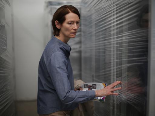 Tilda Swinton steht in einem blauen Hemd zwischen mit Folie verpackten Fenstern.