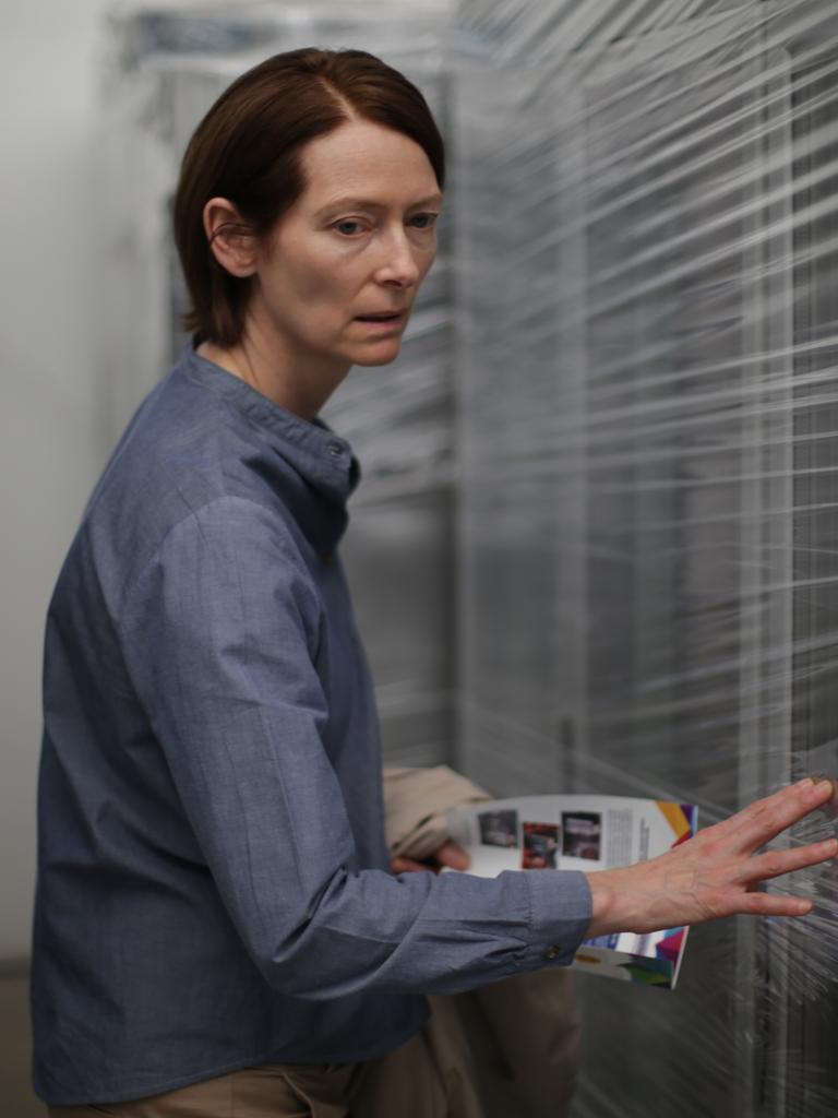 Tilda Swinton steht in einem blauen Hemd zwischen mit Folie verpackten Fenstern.