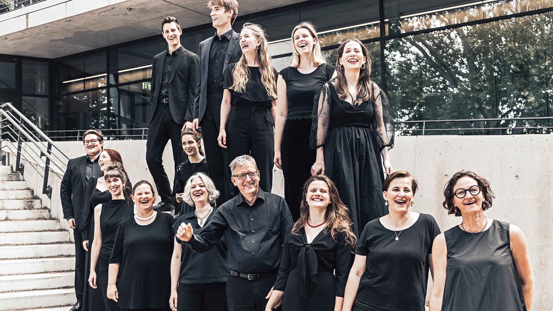 Der Leiter Hans-Christoph Rademann steht von seinem Chor umringt auf einer Außenterrasse mit Treppe, während alle gemeinsam lachen. 