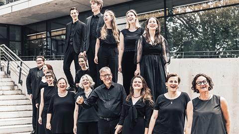 Der Leiter Hans-Christoph Rademann steht von seinem Chor umringt auf einer Außenterrasse mit Treppe, während alle gemeinsam lachen. 