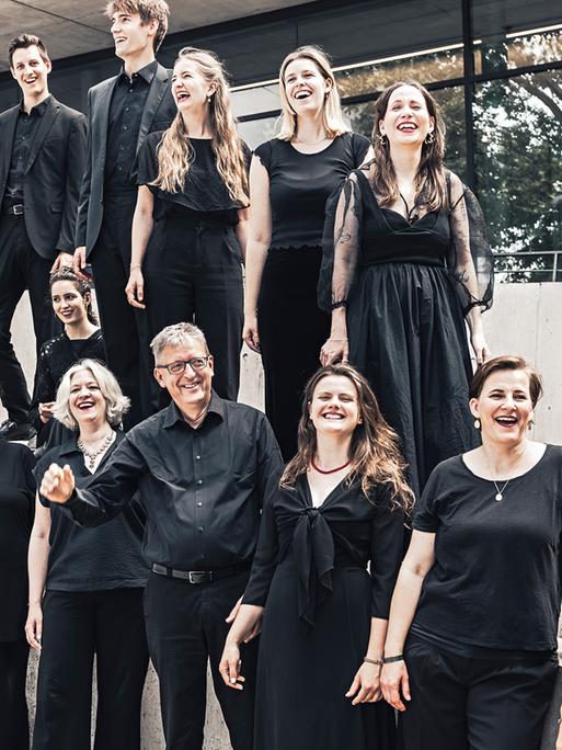 Der Leiter Hans-Christoph Rademann steht von seinem Chor umringt auf einer Außenterrasse mit Treppe, während alle gemeinsam lachen. 