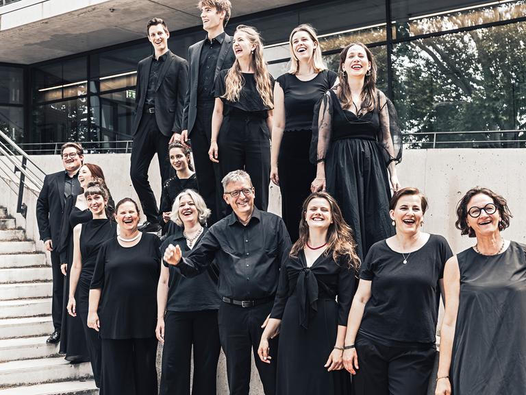 Der Leiter Hans-Christoph Rademann steht von seinem Chor umringt auf einer Außenterrasse mit Treppe, während alle gemeinsam lachen. 