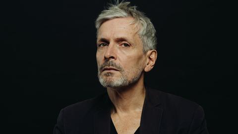 Jan Gerdes stitz mit aufmerksamen Blick vor einem dunklen Hintergrund, sodass sein Gesicht und seine Hände enorm hervortreten.