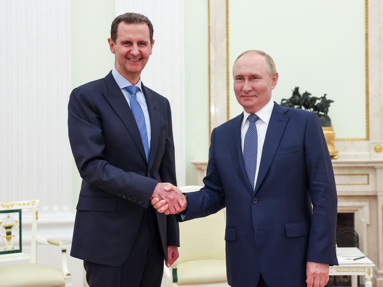 Wladimir Putin und Bashar al-Assad schütteln sich die Hände und lächeln in die Kamera.